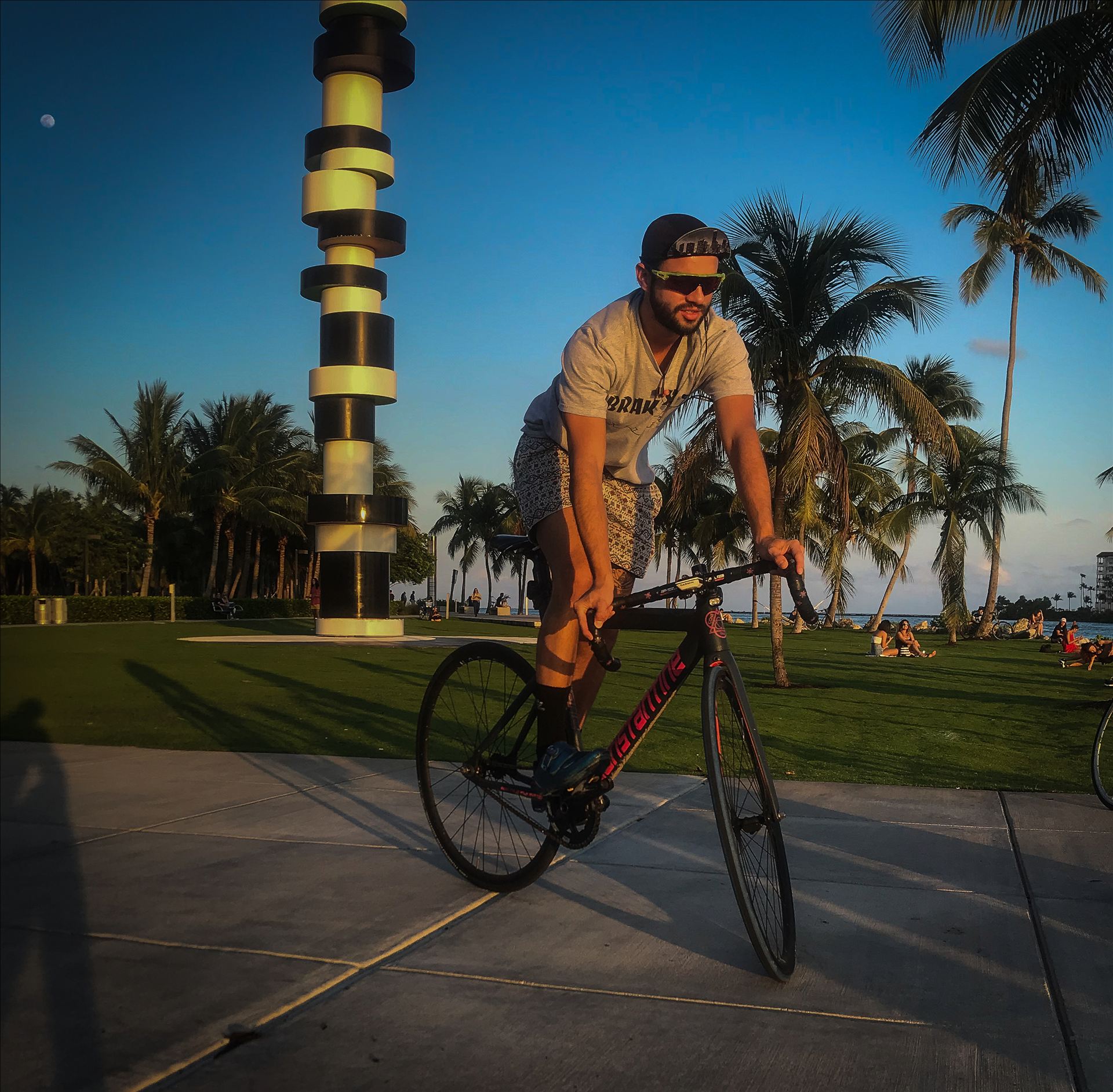 Riding a brakeless fixed-gear bike along the water in Miami at golden hour, palm trees behind.